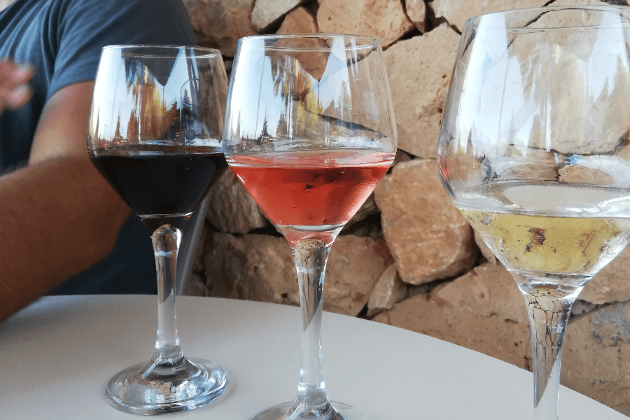 table with three glasses of wines on it: red, rosé and white wine, in front of a stone wall.