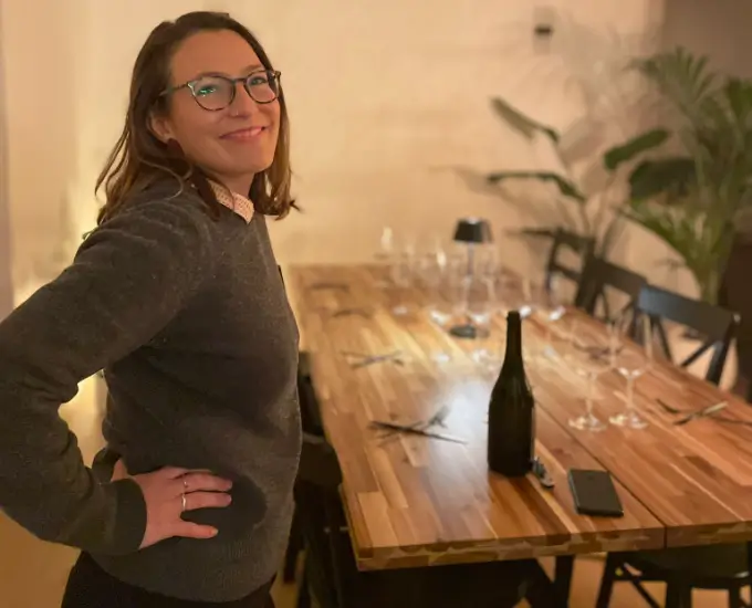 Woman standing next to a table, set for a wine tasting, with glasses and a bottle of wine.